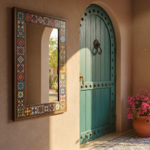 Op de afbeelding: Een rechthoekige spiegel met een houten frame en een kleurrijke tegelrand hangt aan een beige muur. Een turquoise deur met een decoratieve handgreep en een terracotta pot met roze bloemen zijn ook zichtbaar.