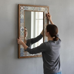 May include: A rectangular mirror with a wooden frame and decorative tile border is being hung on a gray wall. The mirror's frame is a warm brown, and the tile border features a mix of blue, white, and brown patterns.