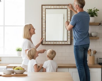 May include: A family hangs a rectangular mirror in a kitchen. The mirror has a brown frame with a decorative border. The family is dressed in casual clothing. The kitchen has white cabinets and a wooden countertop.