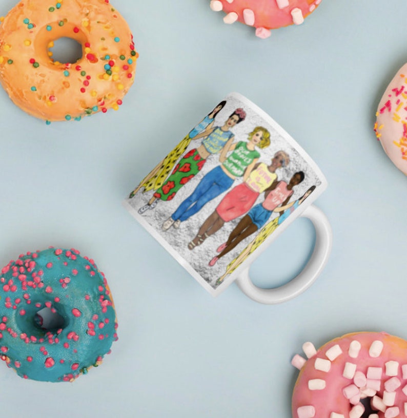 May include: White ceramic mug with a colorful illustration of six women standing together. The women are wearing different outfits and have different hairstyles. The text on the mug reads "Girls Just Wanna Have Fun".