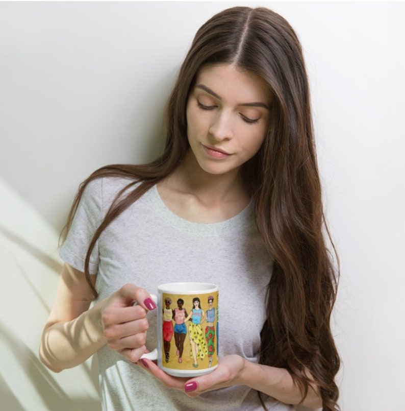 May include: White ceramic mug with a colorful illustration of five women wearing different outfits. The women are standing in a line and smiling. The mug is being held by a person.