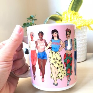 May include: A pink ceramic mug with a graphic of five women walking together. The women are wearing different outfits and have different skin tones. The text on the mug reads "Time's Up" and "United".