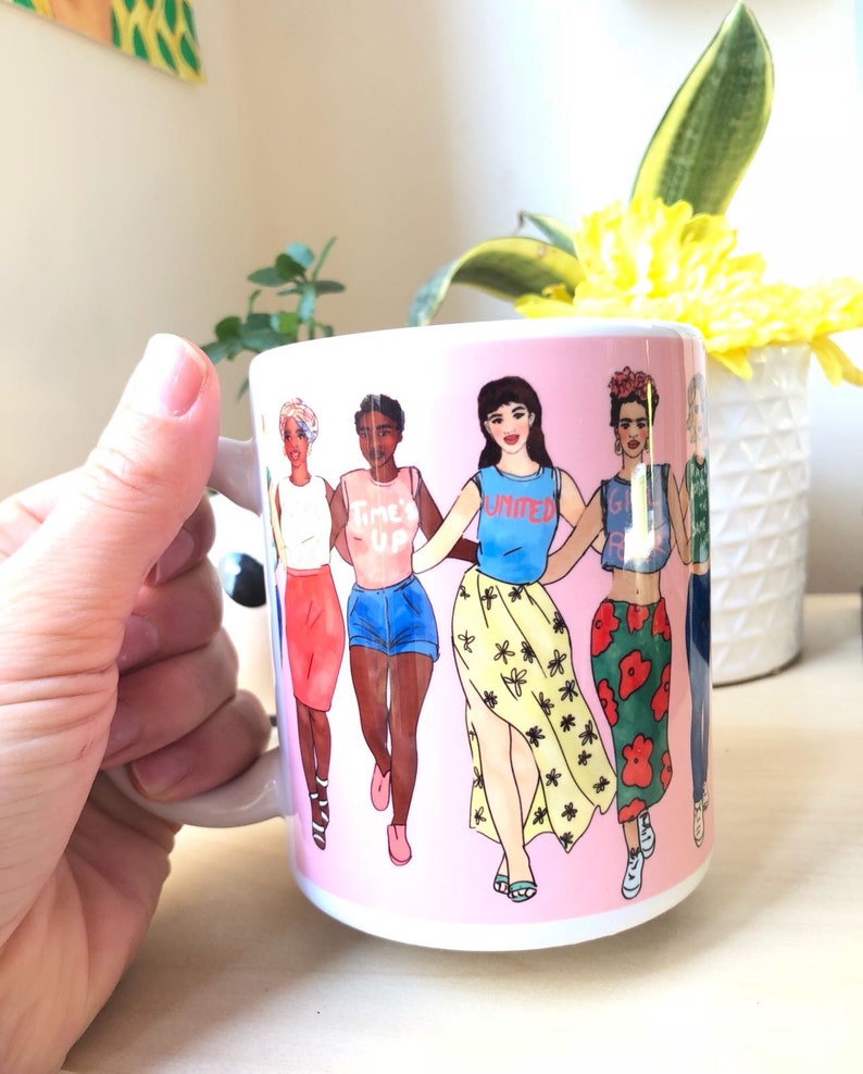 May include: A pink ceramic mug with a graphic of five women walking together. The women are wearing different outfits and have different skin tones. The text on the mug reads "Time's Up" and "United".