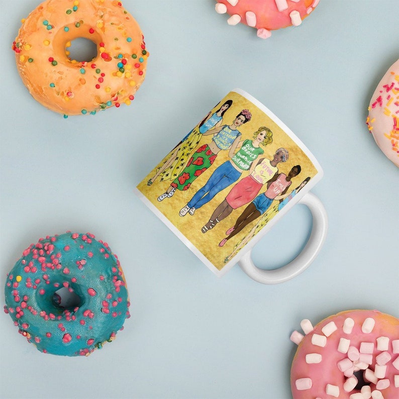 May include: White ceramic mug with a yellow background featuring a group of six women with different skin tones and hairstyles. The women are wearing colorful clothing and holding signs with empowering messages. The text on the signs reads "Our Bodies Our Choices", "Feminist AF", "Trust Me I'm a Doctor", "Girl Power", "Strong Women", and "I'm With Her".