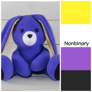 May include: A soft, purple bunny toy with black eyes and a white snout. The bunny's ears have yellow detailing. Beside the bunny is the nonbinary pride flag and the word "Nonbinary".