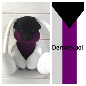 May include: A handmade plush toy with a black head, purple face, and white body. The toy is next to a Demisexual pride flag with black, gray, and purple stripes. The text "Demsexual" is written on the flag.