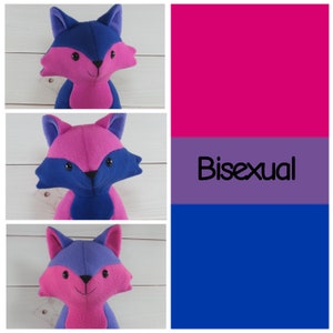 May include: Three plush fox toys with different coloured fur. The first fox has blue ears and a pink body. The second fox has pink ears and a blue body. The third fox has purple ears and a pink body. The background is a bisexual pride flag with the word "Bisexual" written in black.
