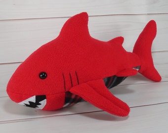 May include: Red plush shark toy with black eyes and a white mouth. The shark has a plaid pattern on its underside.