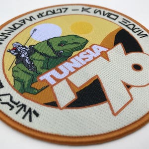 May include: A round, beige patch with an orange border. The patch features a green creature with a rider, a white and yellow sun, and the text "TUNISIA 79" in white. The text "INSIDE EDGE - K.W.D.O.T.V - VINNO7M" is written in black around the border.