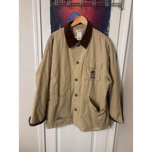 May include: Tan work jacket with a brown corduroy collar and cuffs. The jacket has a button-up front, two front pockets, and a small pocket on the chest. A small logo is visible on the chest pocket. The jacket is hanging on a wooden hanger.