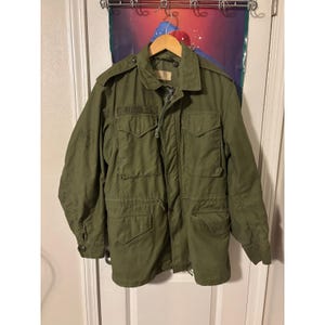 May include: An olive green field jacket featuring a collared neckline, shoulder epaulets, and four large front pockets with buttoned flaps. The jacket includes a central zipper closure, a drawstring waist, and long sleeves with adjustable buttoned cuffs.