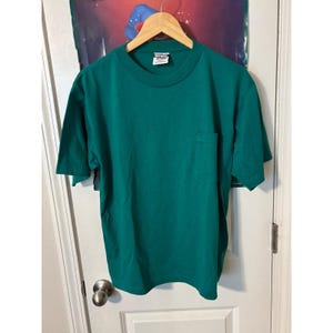 以下が含まれることがあります： 胸ポケットと半袖が付いた、無地のティールグリーンのTシャツ。白いドアを背景に、木製のハンガーに吊るされています。シャツの首元にはタグが付いています。