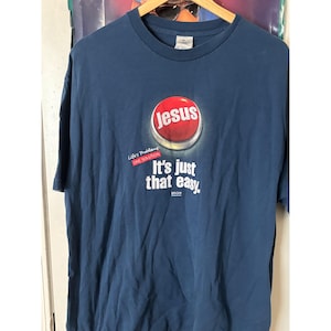 May include: Navy blue t-shirt with a graphic featuring a red button with the word "Jesus" in white. Below the button, the text reads "It's just that easy." The shirt also has the text "Life's Problems, One Solution."