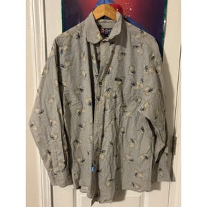 May include: A long-sleeved button-down shirt with a light gray background and a floral pattern in beige and navy blue. The shirt has a collar and a small logo patch. The brand name "Chaps" is visible on the collar.