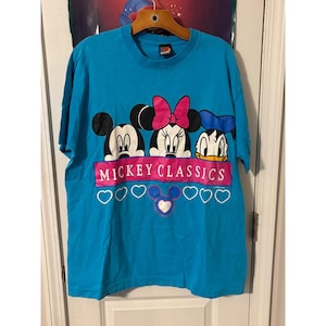May include: A turquoise blue t-shirt featuring Mickey Mouse, Minnie Mouse, and Donald Duck. The shirt has the text "MICKEY CLASSICS" in pink, with heart graphics below. The shirt is hanging on a wooden hanger.