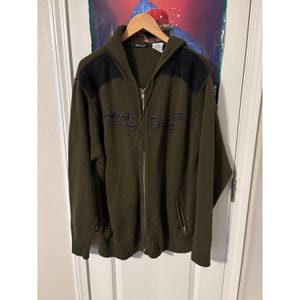 Puede incluir: Sudadera con capucha y cremallera verde oliva con parches negros en los hombros. La sudadera presenta el texto bordado "Blind City" en el pecho. La sudadera está colgada en una percha de madera.