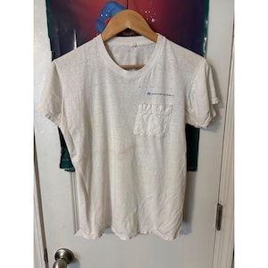 Puede incluir: Una camiseta blanca de manga corta con cuello redondo y un bolsillo en el pecho. La prenda presenta un pequeño logotipo azul y el texto Boardworks impreso sobre el bolsillo. El tejido parece ligero y ligeramente translúcido.