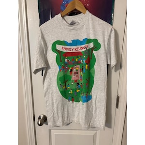 Può includere: T-shirt grigio chiaro con una grafica colorata di una riunione di famiglia. Il design include uno striscione con la scritta "FAMILY REUNION" e raffigura persone che svolgono varie attività in un ambiente di parco. La maglietta ha maniche corte e girocollo.