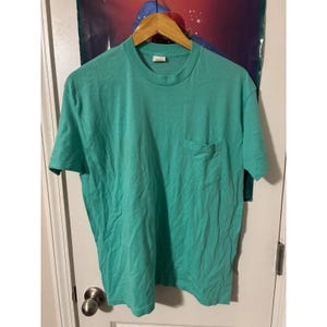 May include: A teal short-sleeved t-shirt with a chest pocket, hanging on a wooden hanger. The fabric has a slightly wrinkled appearance. The shirt is a solid colour and features a crew neck. The shirt is a casual wear item.