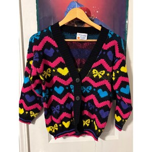 以下が含まれることがあります： ピンクのジグザグ、青いハート、黄色のリボン、紫のハートのカラフルなパターンが特徴の、Vネックのヴィンテージブラックカーディガン。セーターには3つの黒いボタンがあり、木製のハンガーに吊るされています。
