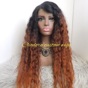May include: A long, wavy wig with a black top that fades into a reddish-brown color. The wig is styled on a mannequin head with a dark complexion, wearing a black and gold necklace. The text "Cleodora custom wigs" is visible.