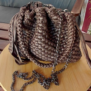 May include: A metallic bronze-colored crocheted bucket bag with a drawstring closure. The bag features a thick, braided handle made of the same material and a detachable dark metal chain shoulder strap. The texture is dense and uniform.