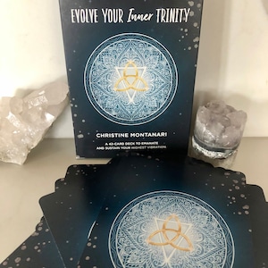 Puede incluir: Una caja azul con un diseño de mandala dorado y blanco y el texto "Evolve Your Inner Trinity" y "Christine Montanari" con el texto "Una baraja de 42 cartas para emanar y sostener tu vibración más alta."