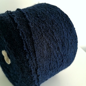 May include: A large, cylindrical spool of dark navy blue yarn with a highly textured, nubby surface. The yarn is tightly wound around a central white cardboard core, showing a consistent deep blue color throughout the entire length of the fiber.