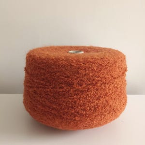 May include: A large spool of orange yarn with a textured, fuzzy surface. The yarn is cylindrical with a silver core. The spool is sitting on a white surface against a plain white background. This yarn is suitable for knitting and crochet projects.