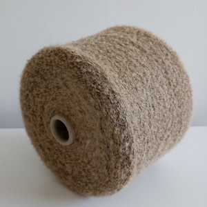 May include: A large spool of yarn, primarily beige with darker brown flecks, is displayed. The yarn is tightly wound, creating a textured surface. The core of the spool is visible, featuring a light-colored cardboard tube. Ideal for knitting and crochet projects.