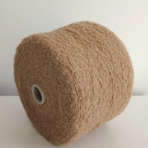 May include: A large spool of light brown, textured bouclé yarn. The yarn features a fuzzy, looped surface and is wound tightly around a central white cardboard core, creating a cylindrical shape with a soft, consistent tan color throughout.