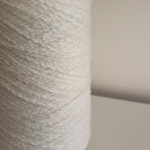 May include: A large cylindrical spool of off-white yarn featuring a textured, slightly crinkled appearance. The thread is tightly wound in a consistent pattern around the central core, creating a uniform surface with visible fibre strands.