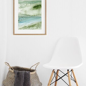 May include: A framed abstract painting with green and blue watercolor washes. The painting is hanging on a white wall above a white chair and a woven basket.