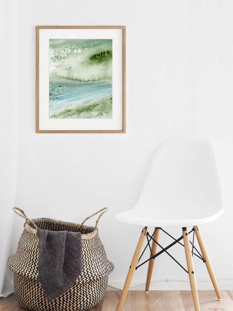 May include: A framed abstract painting with green and blue watercolor washes. The painting is hanging on a white wall above a white chair and a woven basket.