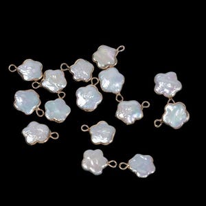 Puede incluir: Una colección de abalorios de perlas blancas en forma de pétalo con bordes de metal dorado y pequeños bucles para sujetar a la joyería. Las perlas tienen un brillo iridiscente y se colocan sobre un fondo negro.