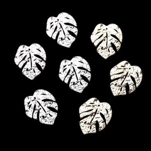 Puede incluir: Siete colgantes blancos con forma de hoja de monstera, texturizados y con agujero para ensartar. Los colgantes están dispuestos en un patrón circular sobre un fondo negro. Las hojas tienen un aspecto moteado y envejecido.