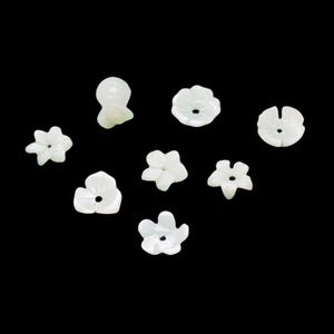 Könnte beinhalten: Eine Sammlung weißer, blütenförmiger Perlen mit unterschiedlichen Blütenblattdesigns. Die Perlen bestehen aus einem glänzenden Material und haben ein Loch in der Mitte zum Auffädeln. Die Perlen sind auf einem schwarzen Hintergrund verstreut.