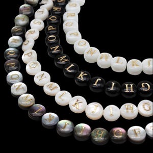 Op de afbeelding: Close-up van verschillende strengen kralen met letters in wit, zwart en iriserende kleuren. De gouden letters zijn zichtbaar op de kralen. De kralen zijn gerangschikt in een gebogen patroon tegen een zwarte achtergrond. De kralen zijn waarschijnlijk voor het maken van sieraden.
