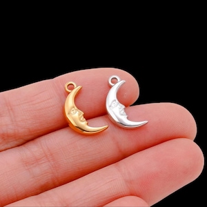 Könnte beinhalten: Zwei halbmondförmige Anhänger, einer goldfarben und einer silberfarben, jeweils mit detailliertem Gesichtsdesign. Die Anhänger haben oben eine kleine Schlaufe zum Befestigen. Der Hintergrund ist schwarz.