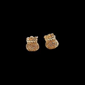 May include: A pair of gold-toned stud earrings shaped like small drawstring pouches. Each earring features a detailed, intricate filigree pattern across the surface with a gathered top edge, creating a textured, metallic appearance against a black background.
