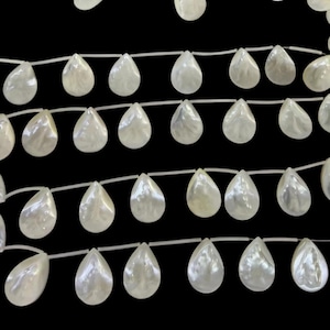 May include: A strand of white, teardrop-shaped mother-of-pearl beads arranged in multiple horizontal rows against a black background. Each bead features a smooth, iridescent surface with a subtle luster and a central hole for stringing.