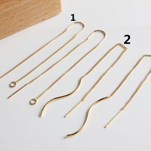 Può includere: Orecchini a filo dorati in vari design. L'immagine mostra tre paia di orecchini, ciascuno con catene delicate e barre curve o dritte. Gli orecchini sono esposti su una superficie bianca.