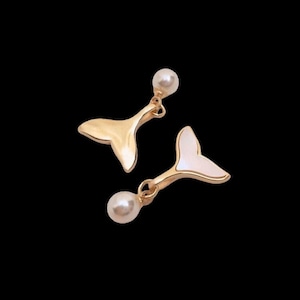 Może przedstawiać: Para złotych kolczyków z motywem ogona syreny. Każdy kolczyk ma małą, okrągłą, perłową koralik na górze i zawieszkę w kształcie ogona. Jeden ogon jest złoty, drugi biały.