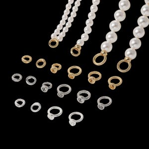 May include: A collection of jewelry components featuring four strands of white faux pearls with gold-toned circular end connectors, accompanied by an assortment of small gold-toned and silver-toned metal rings, some with textured edges and settings.