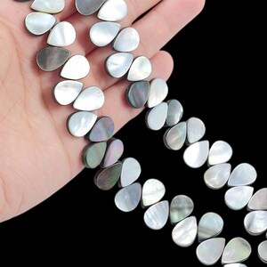 May include: A strand of teardrop-shaped beads crafted from iridescent mother-of-pearl shell. The beads feature a smooth, polished surface with a natural color palette ranging from creamy white to dark gray, showcasing a distinct pearlescent luster.