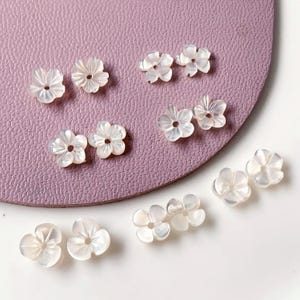 Può includere: Una collezione di piccole perline bianche a forma di fiore. Le perline hanno una lucentezza perlescente e un foro al centro. Sono disposte su una superficie viola chiaro e una superficie bianca.