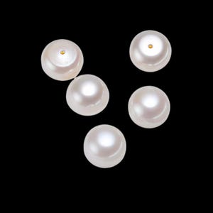 May include: Five lustrous, round pearls against a black background. The pearls have a creamy white color with a subtle sheen. Each pearl has a small, golden accent on its surface, adding a touch of elegance.