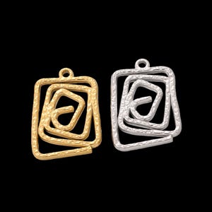 May include: Two metal pendants, one gold and one silver, each with a square frame and an inner spiral design. The pendants have a textured surface and a small loop at the top for hanging. The background is black.