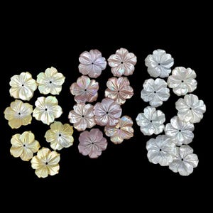 May include: A collection of flower-shaped mother-of-pearl beads in various colors, including white, pale pink, and yellow. The beads have a carved petal design and a central hole. The background is black.