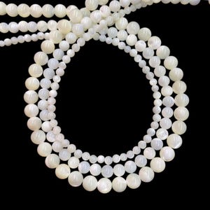 May include: A collection of smooth, round, off-white mother of pearl beads arranged in multiple concentric circular strands. The beads vary in size, featuring a lustrous, iridescent finish that reflects light against a solid black background.